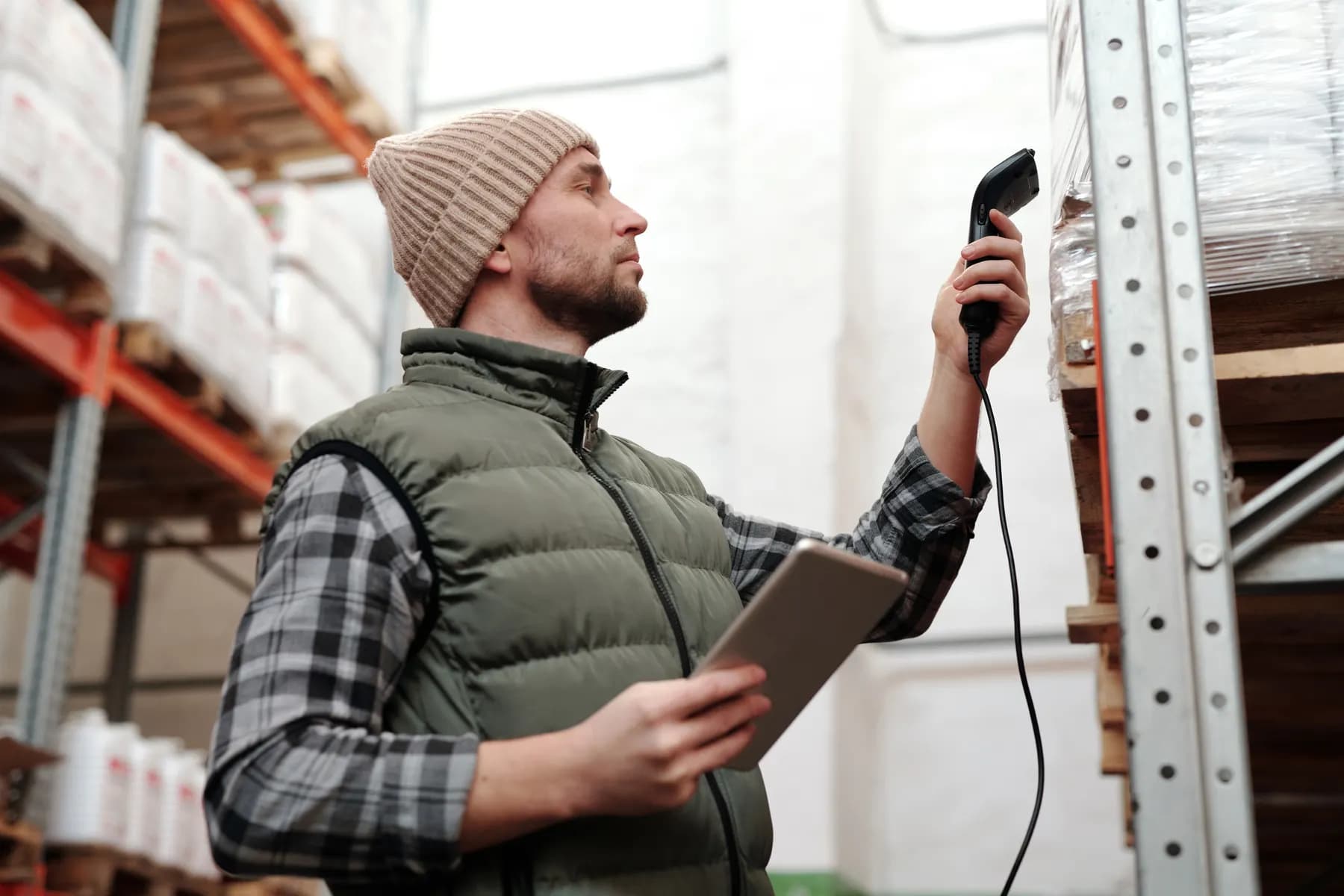 A warehouse worker scans a packaged product on a shelf while holding a tablet.