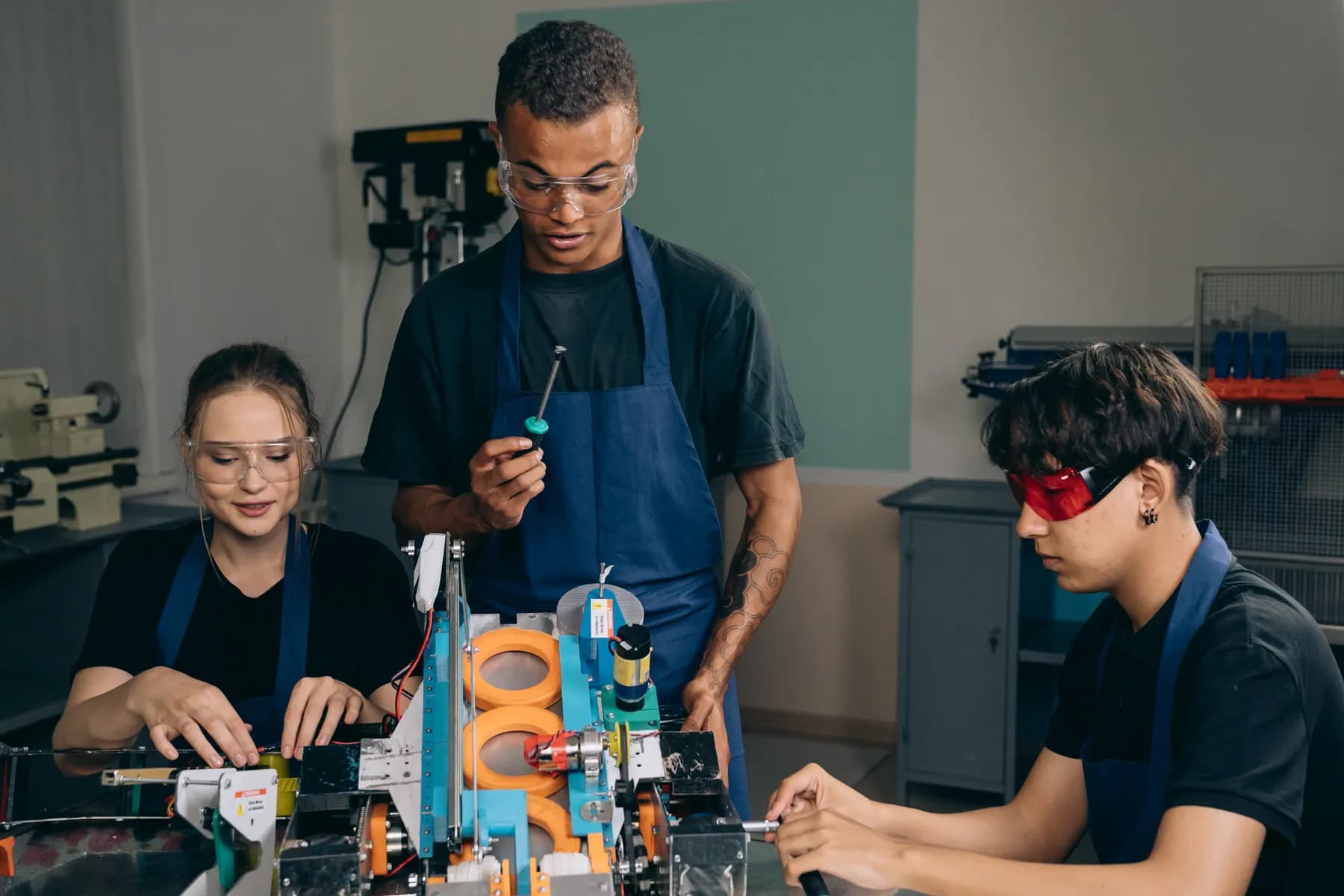 Three people wearing safety glasses work together on a machine in a workshop.